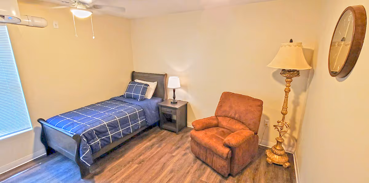 A small bedroom with a single bed covered in blue and white checkered bedding, a wooden nightstand with a lamp, a brown recliner chair, a tall ornate floor lamp, a wall clock, and a window with closed blinds.