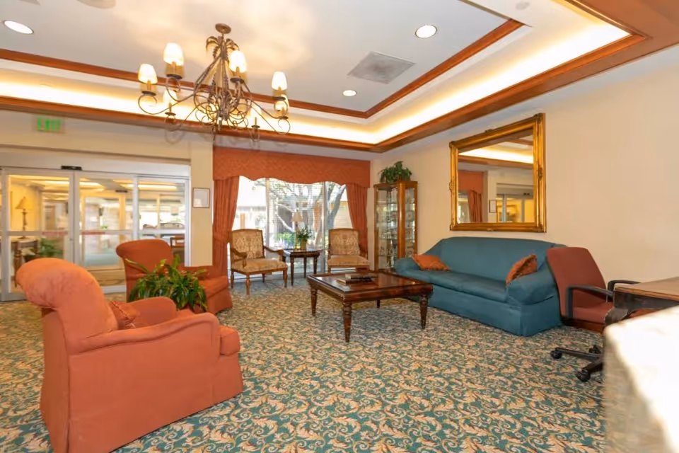 A cozy living room area with a patterned carpet, a teal sofa with two orange pillows, two orange armchairs, two wooden chairs with patterned cushions, a wooden coffee table, a large gold-framed mirror on the wall, a chandelier hanging from the ceiling, and a glass display cabinet near a window with orange curtains. The room has recessed lighting and a view of an outdoor area through glass doors.