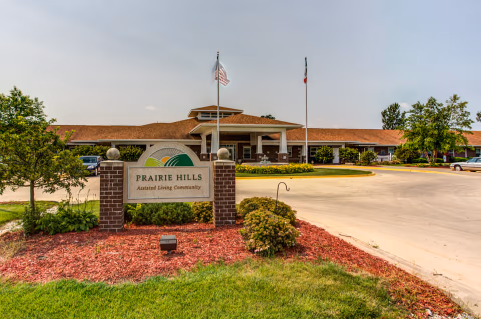 Exterior view of Prairie Hills Assisted Living Community building with a sign in front, two flagpoles with flags, landscaped bushes, and a driveway.