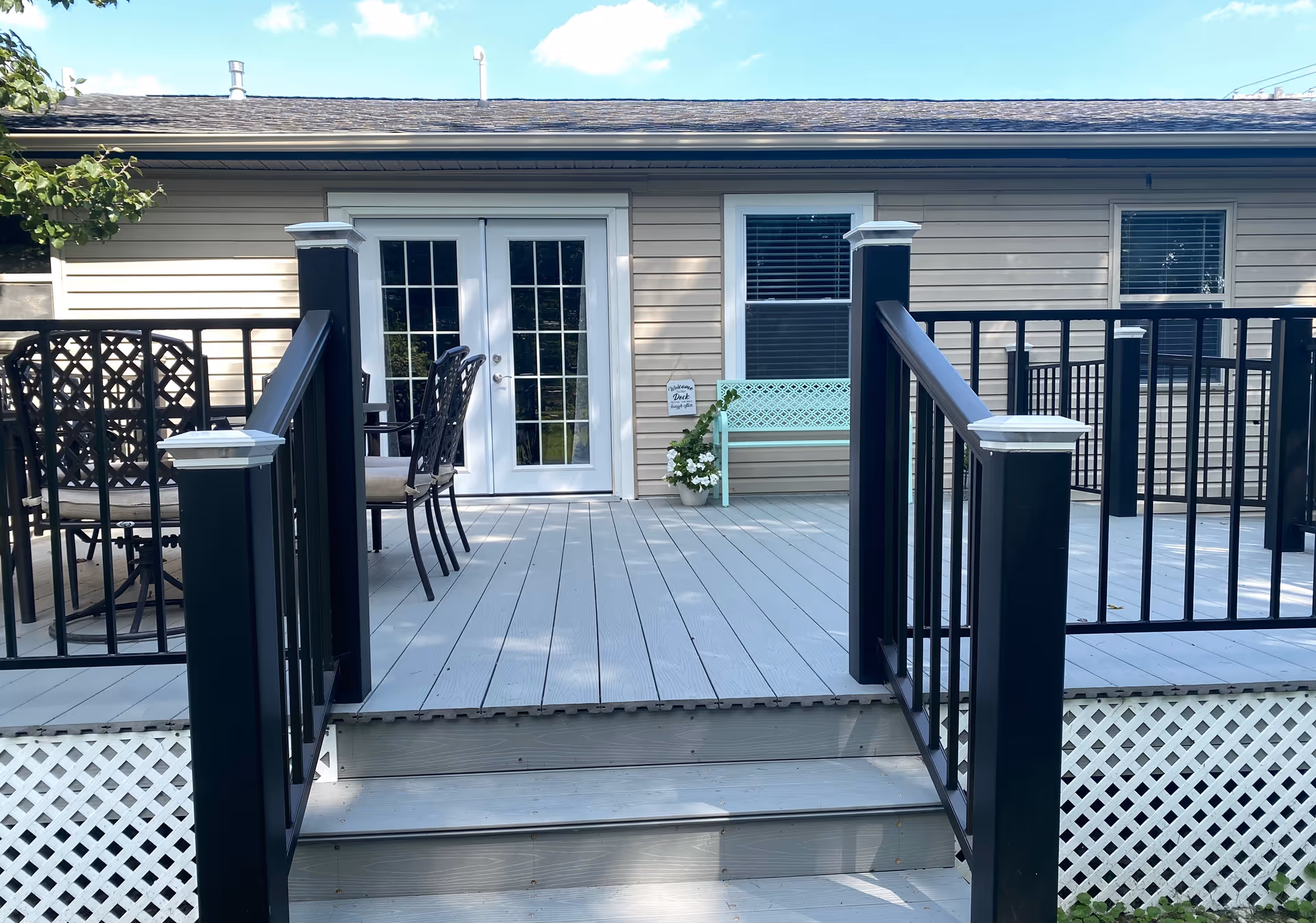 Outdoor deck area with black railings and steps leading up to it. There is a table with chairs on the left side and a mint green bench with a potted plant and a small sign on the wall near a window. The deck is attached to a beige house with white-framed French doors and windows.