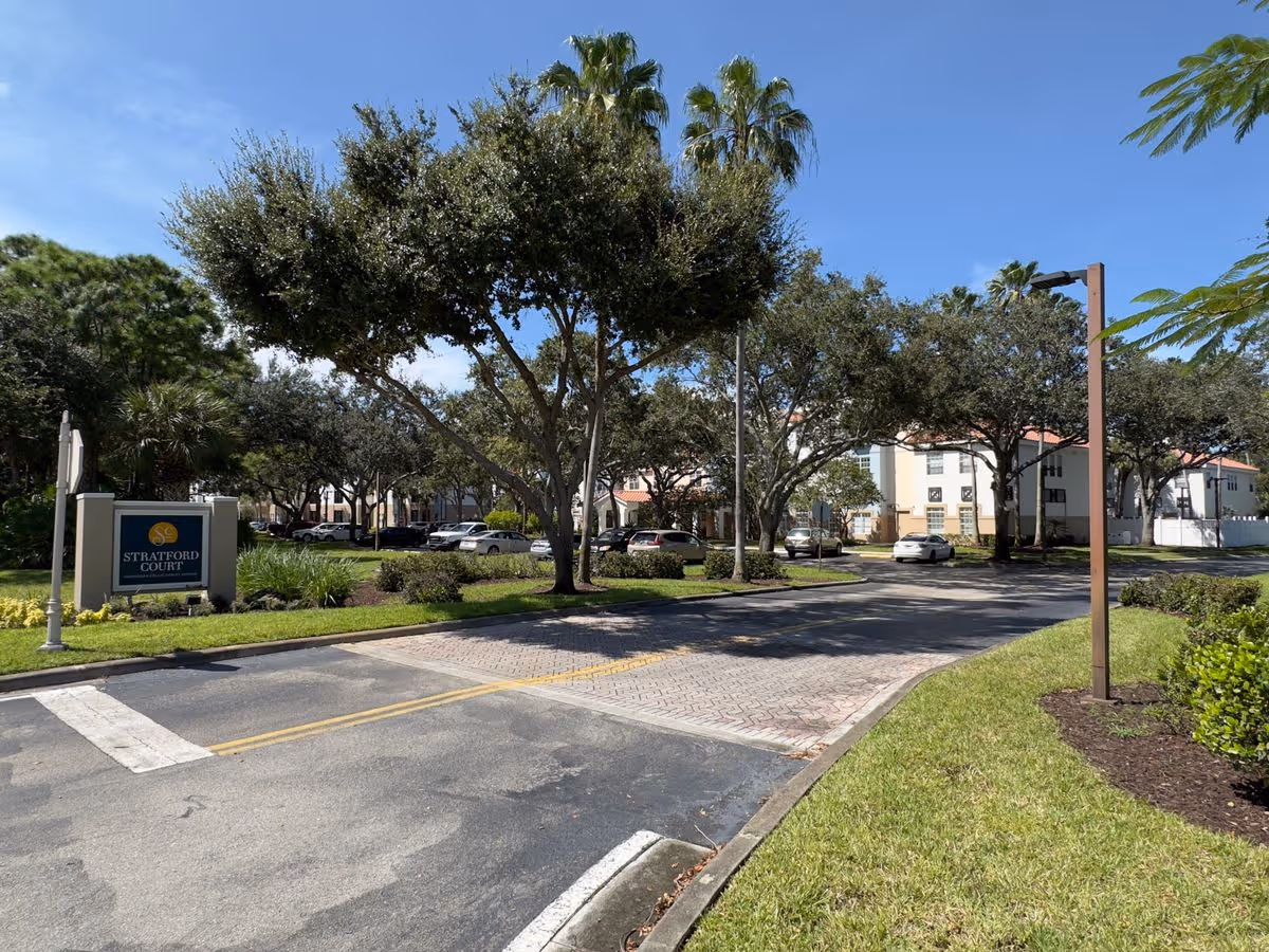 Entrance driveway and landscaped grounds in front of a multi-story residential complex with a sign reading 'Stratford Court' and parked cars under trees.