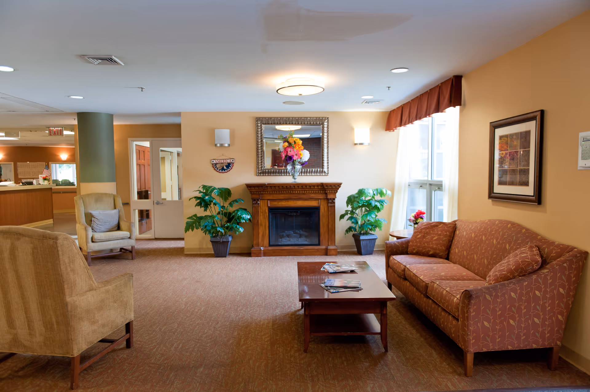 Comfortable sitting area with sofas, armchairs, a coffee table, fireplace, plants, and a mirror in a senior living facility.
