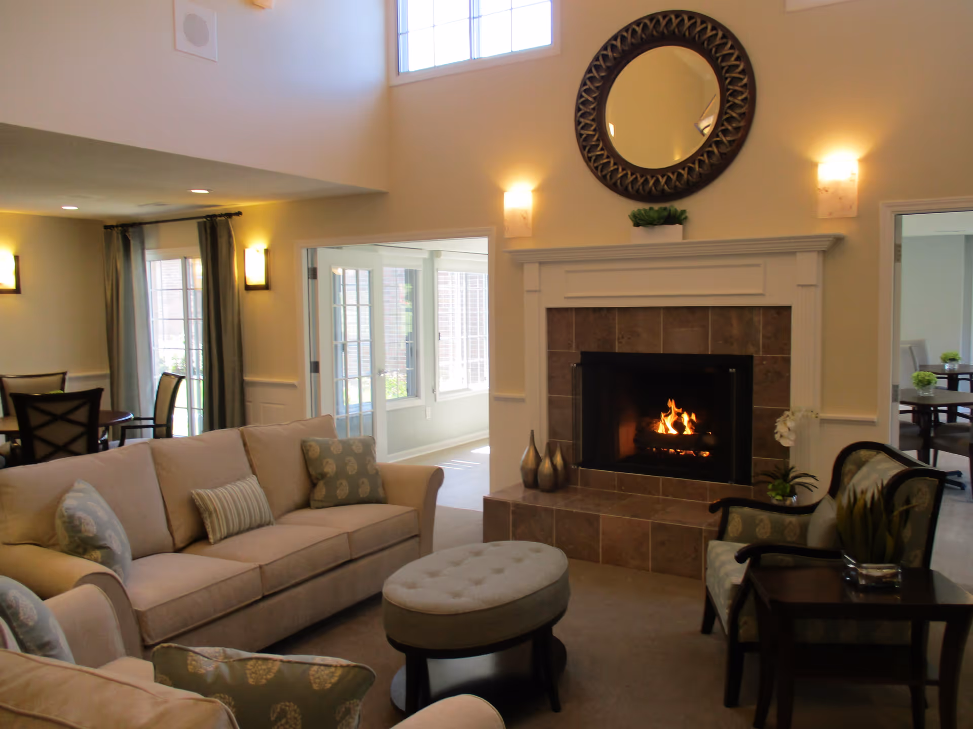 A cozy living room with a beige sofa, patterned cushions, an upholstered ottoman, two armchairs, and a fireplace with a fire burning. Above the fireplace is a decorative round mirror. The room has high ceilings and windows letting in natural light. Adjacent rooms with dining tables and chairs are visible through open doorways.