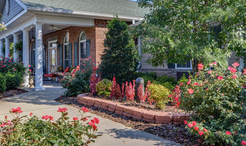 Front exterior view of a brick building with white columns and a covered porch. The porch has seating with red cushions. There are well-maintained flower beds with pink and red flowers and green shrubs along a concrete walkway leading to the entrance.