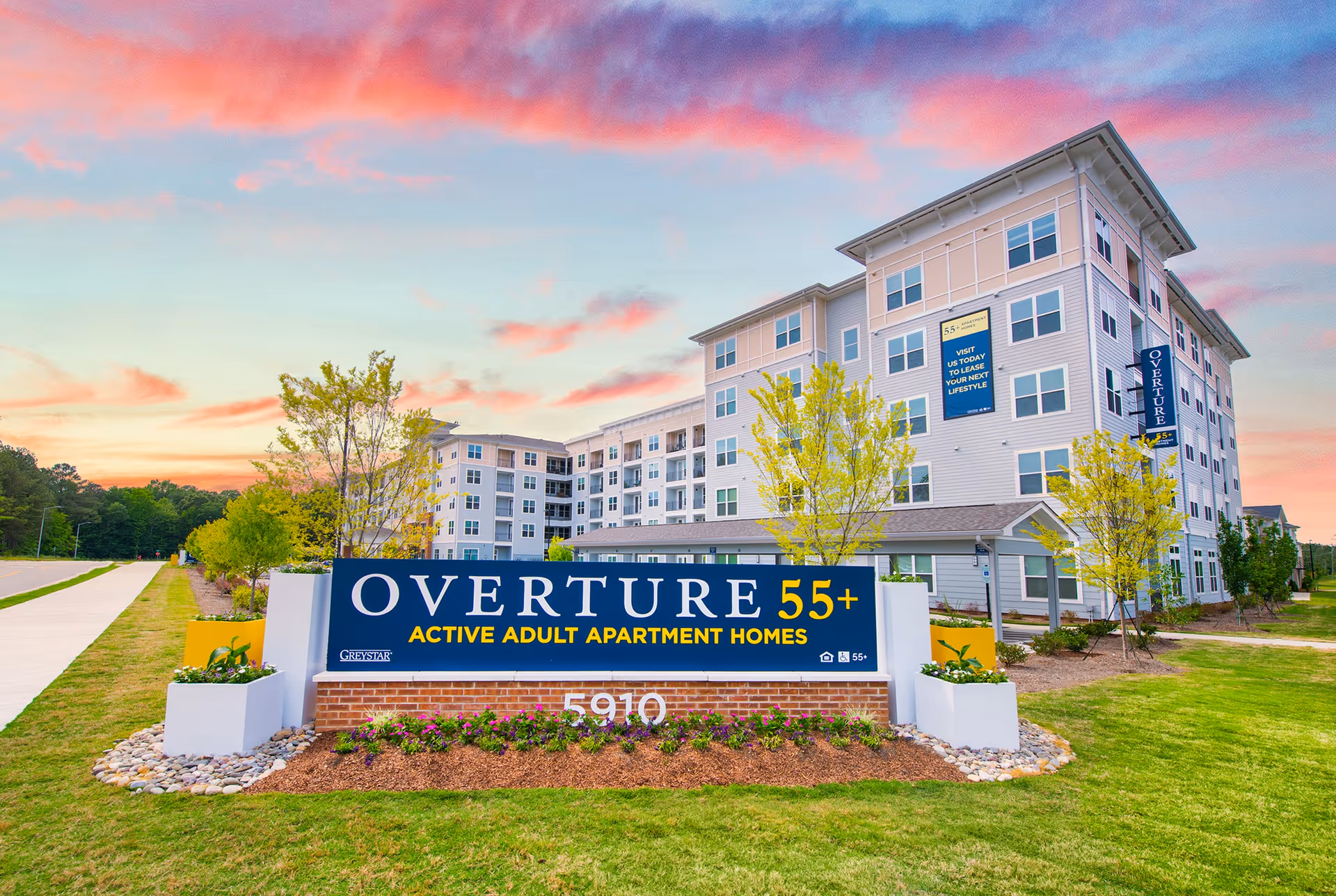 A large Overture 55+ active adult apartment building with a prominent entrance sign on landscaped grounds under a colorful sky.