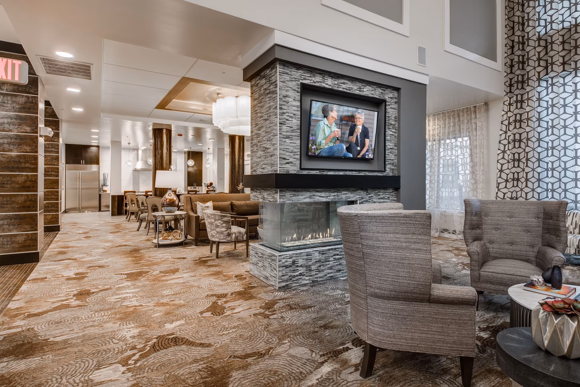 A modern and spacious senior living facility common area with a cozy fireplace surrounded by gray tile, a flat-screen TV mounted above it showing two elderly women talking. The room features comfortable seating including gray armchairs and a sofa, patterned carpet, large windows with sheer and patterned curtains, and a dining area with tables and chairs in the background near a kitchen area with stainless steel appliances.