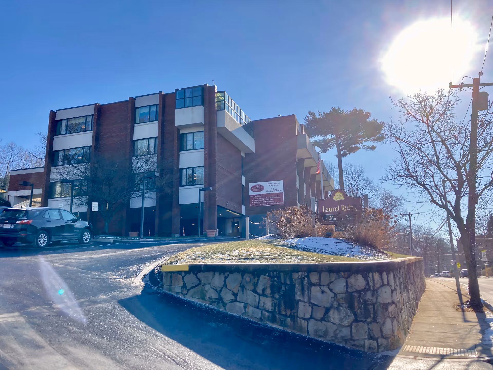 Exterior view of a multi-story brick building with large windows, identified as Laurel Ridge Rehab And Skilled Care Center. The building is situated on a slight hill with a stone retaining wall and a sign in front. There is a car parked on the driveway, leafless trees, and a bright sun in a clear blue sky.