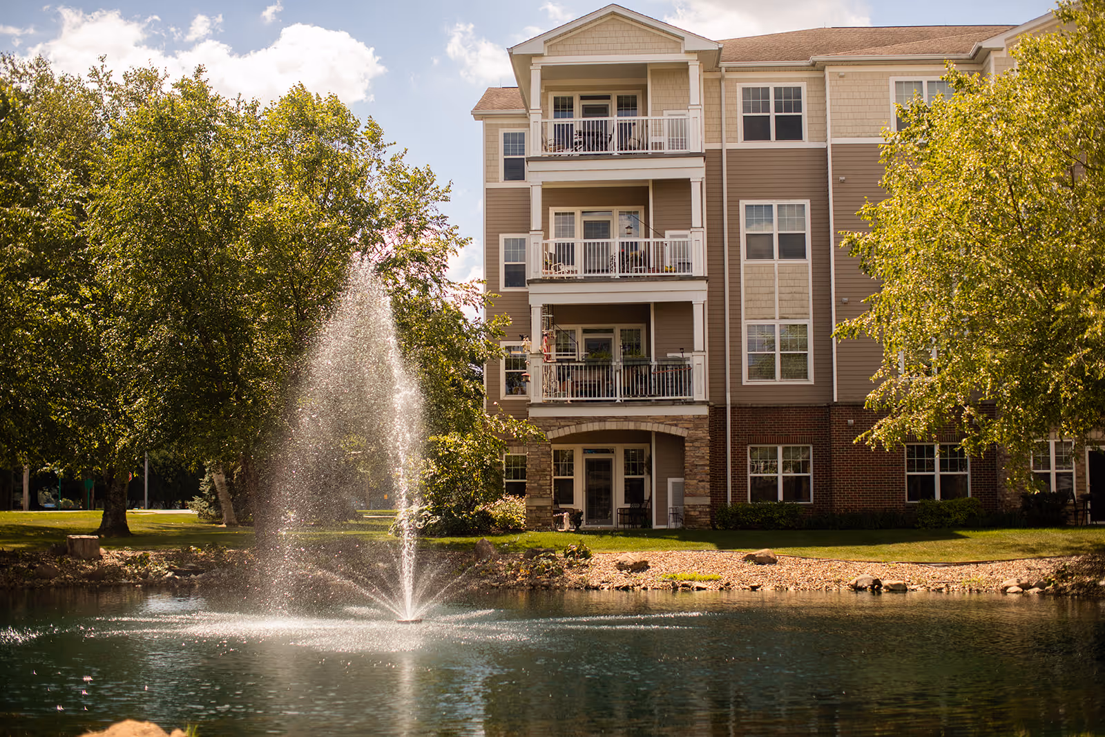 A multi-story residential building with balconies overlooking a pond with a water fountain in the foreground, surrounded by green trees and a grassy area under a partly cloudy sky.