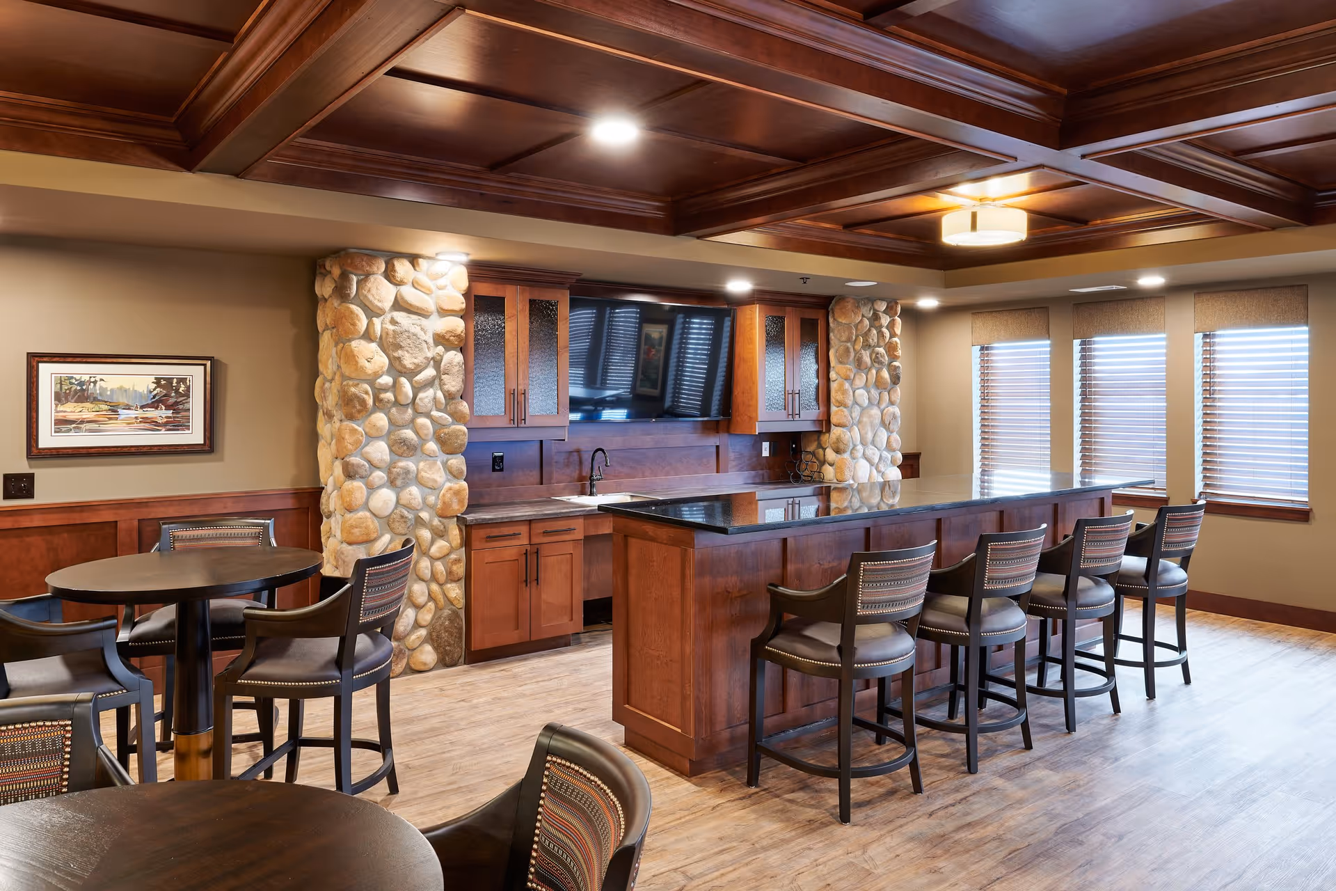 A cozy common area with a long wooden counter and six high chairs, stone pillars, wooden cabinets with a mounted flat-screen TV, round tables with chairs, and windows with blinds letting in natural light.
