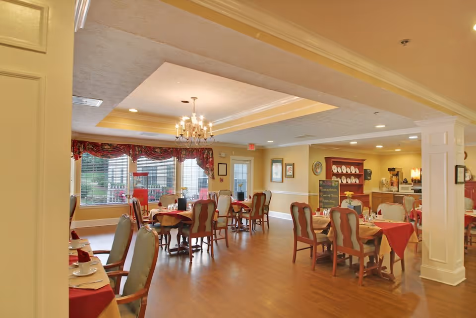 A dining room in a senior living facility with several tables covered with red and beige tablecloths, each set with cups, plates, and napkins. The room has wooden flooring, a chandelier hanging from a recessed ceiling, large windows with floral curtains, and a buffet area with plates and a coffee machine in the background.