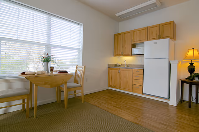 Small apartment kitchenette with wooden cabinets, a white refrigerator and microwave, and a small dining table by a large window.