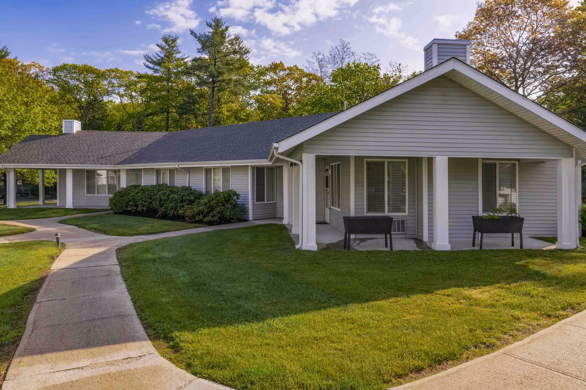 Exterior view of a single-story residential building with gray siding and a dark shingled roof, surrounded by green grass and trees under a blue sky. A concrete pathway curves in front of the building leading to a covered porch area with two black planters.