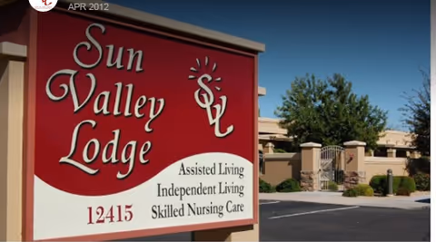 Outdoor view of a large red and white sign for Sun Valley Lodge displaying services including Assisted Living, Independent Living, and Skilled Nursing Care, with the address 12415. In the background, there is a gated entrance with trees and a building under a clear blue sky.