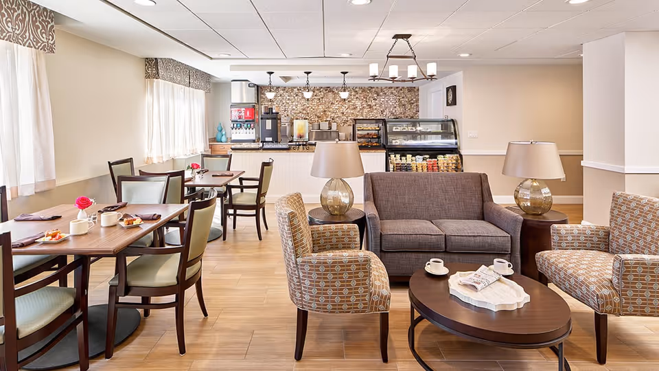 A bright and cozy common area in a senior living facility featuring a small dining section with tables and chairs set with cups and plates, and a lounge area with a gray sofa, two patterned armchairs, a wooden coffee table with cups and a newspaper, and two side tables with lamps. In the background, there is a counter with a coffee machine, beverage dispensers, and a display case with snacks.