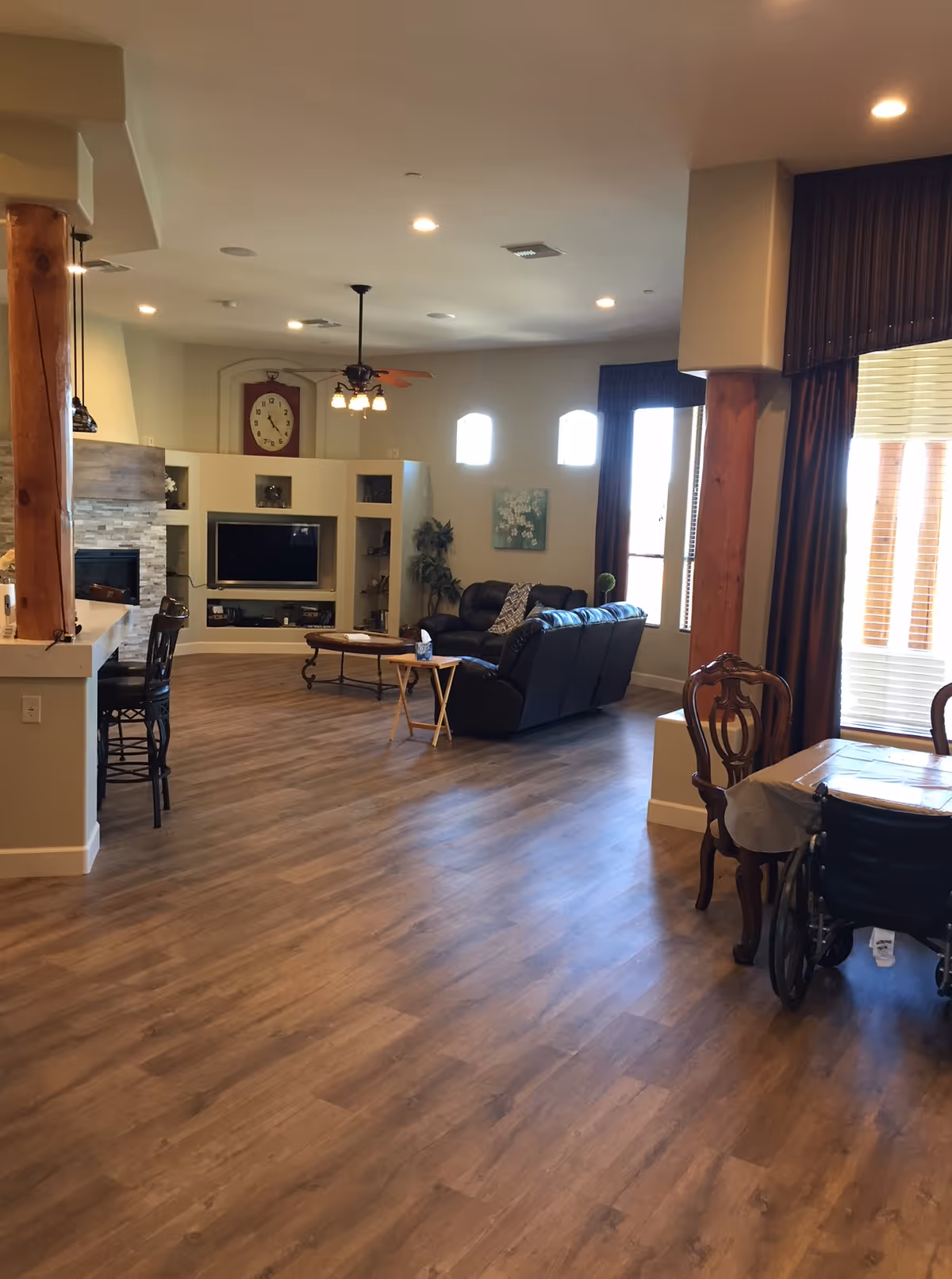 Open, spacious assisted living common area with wood floors, a seating area with leather sofas and TV, dining table and large windows.