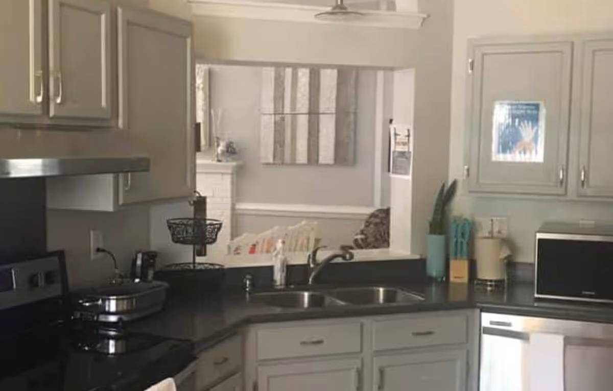 A kitchen area with light gray cabinets, a black countertop, a double sink, a microwave, and a stove. There are kitchen utensils in a holder, a soap dispenser, and a two-tiered basket on the counter. Through a pass-through window, a living room with a white fireplace, a piece of wall art, and a white chair with a cushion is visible.