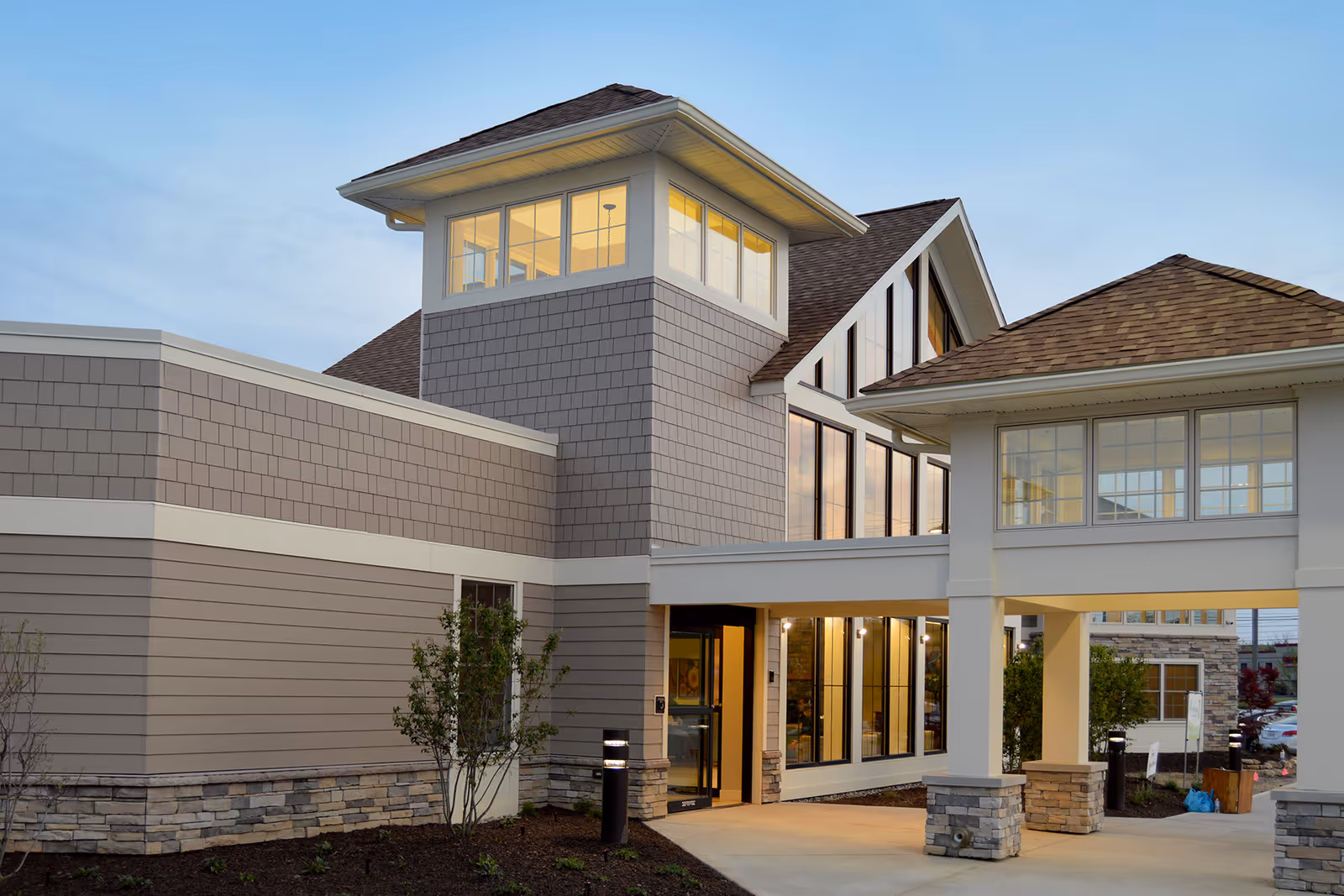 Exterior view of a modern senior living facility building with large windows, stone and siding facade, and a covered entrance walkway during early evening.