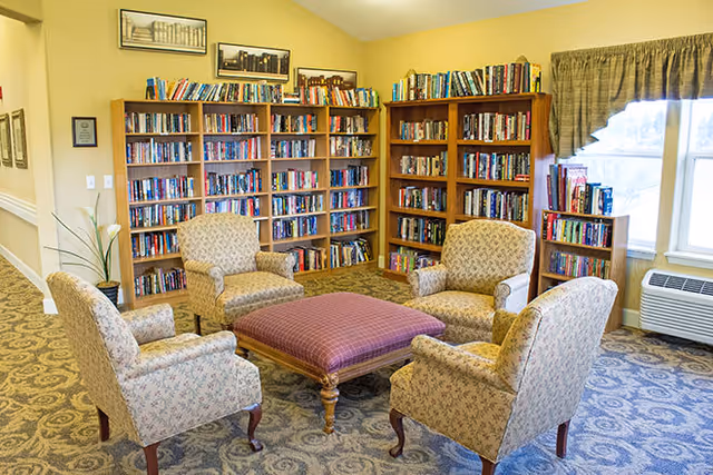 A cozy reading room with four upholstered armchairs arranged around a large square ottoman. Behind the chairs are multiple wooden bookshelves filled with books. A window with a valance lets in natural light, and a small air conditioning unit is below the window. The room has patterned carpet and light-colored walls with framed pictures above the bookshelves.