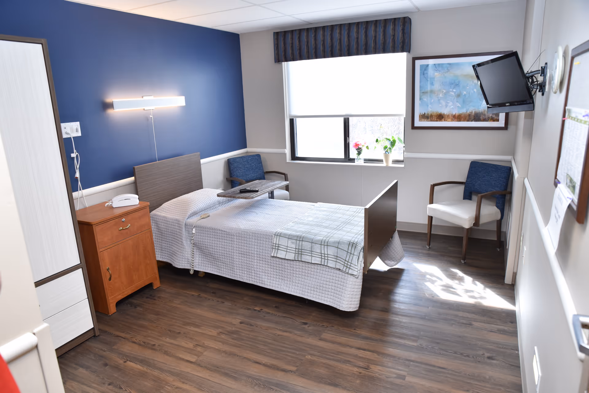 Private senior living bedroom with a single bed, bedside cabinet, two chairs, a window with plants, and a wall-mounted TV.