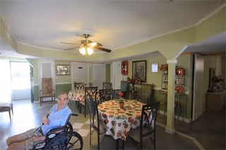 A common area in Mi Casa Adult Living Facility featuring a man in a wheelchair in the foreground. The room has multiple tables covered with floral tablecloths and surrounded by chairs. The walls are painted light green with white trim, and there is a ceiling fan with lights. Various decorations and framed pictures are visible on the walls.