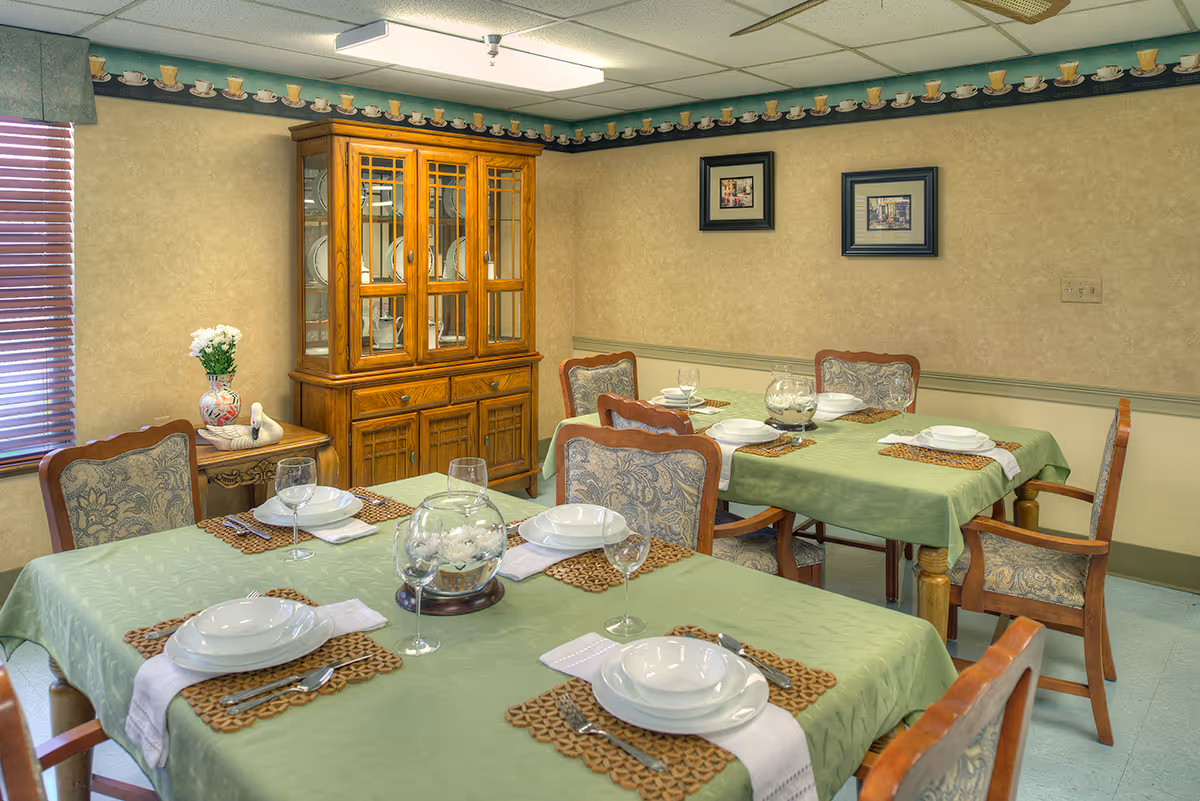 A dining room with two tables covered in green tablecloths, each set with white plates, bowls, wine glasses, and silverware on woven placemats. The room has upholstered wooden chairs, a wooden china cabinet displaying plates, a small side table with a vase of flowers, and framed pictures on the beige walls. A window with wooden blinds is partially visible on the left.