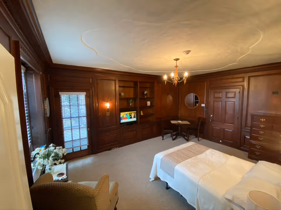 A cozy bedroom with dark wood paneling on the walls, a bed with white linens and a beige throw, a small round table with two chairs, a built-in wooden shelving unit with a small TV, a chandelier hanging from the ceiling, a window with blinds, and a comfortable armchair near a small table with a flower arrangement.