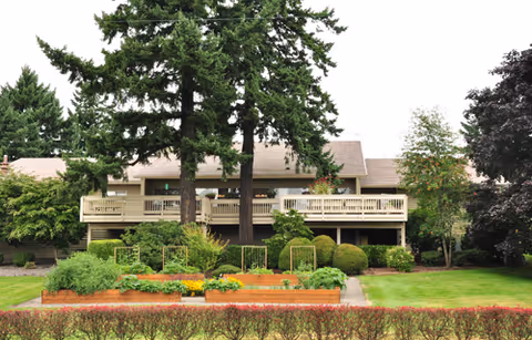 Front exterior of a rehab facility with a two-story building, raised balconies, large evergreen trees, and manicured gardens and lawn.