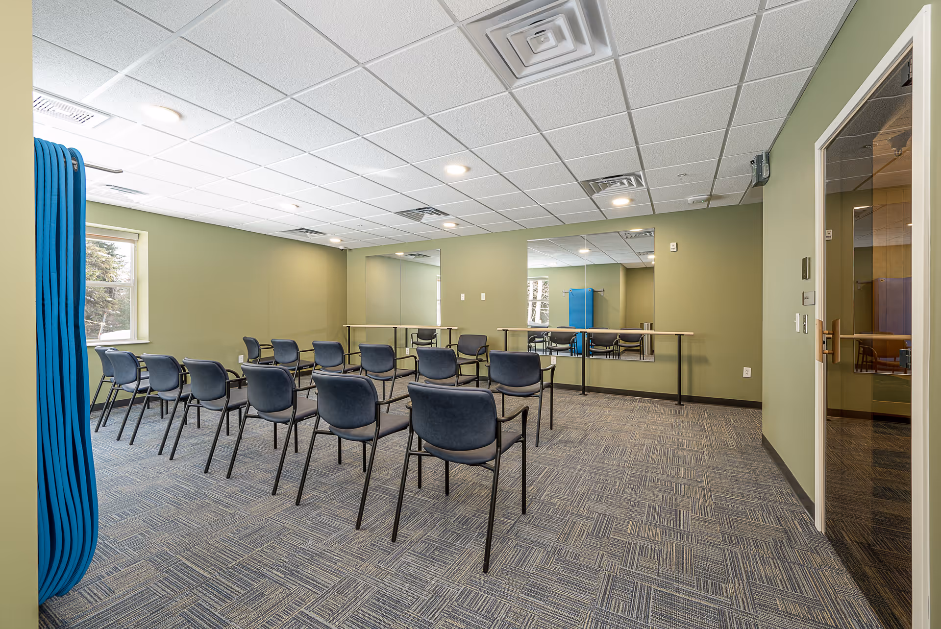 A small meeting/activity room with rows of chairs facing a mirrored wall, green walls, and a carpeted floor.