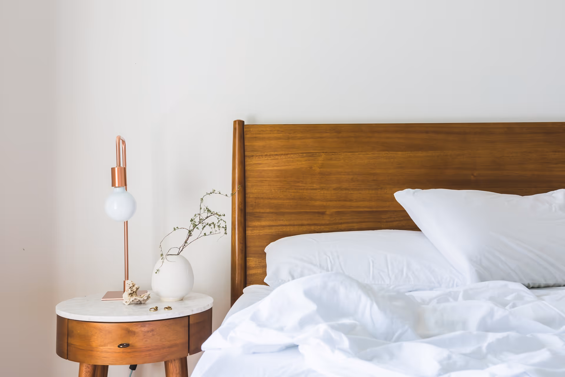 A wooden headboard bed with white linens beside a round bedside table holding a lamp and a small vase.