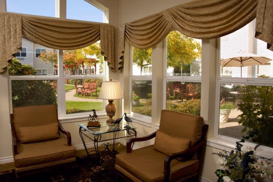 A cozy sitting area with two cushioned armchairs and a glass-top side table with a lamp and decorative items, positioned in front of large windows with beige valance curtains. Outside the windows, there is a garden area with benches, trees, and an umbrella-covered table.