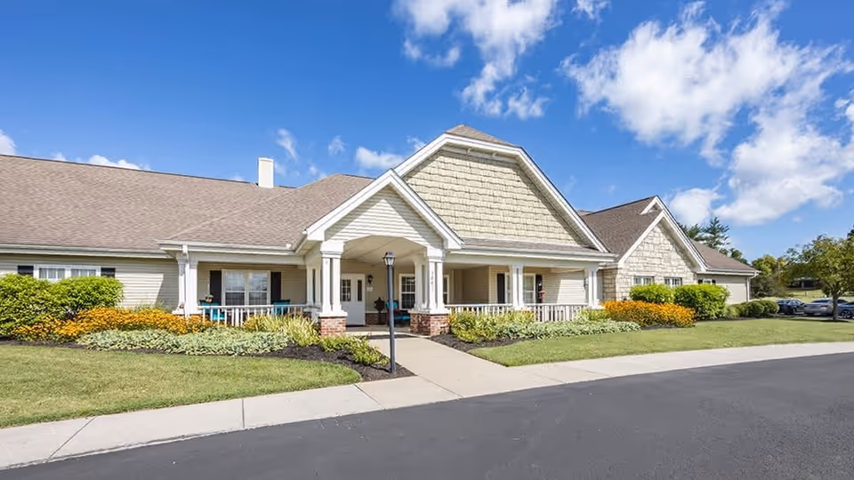 Exterior view of a single-story senior living facility building with beige siding, a covered entrance with white columns, well-maintained landscaping including green bushes and yellow flowers, a paved driveway, and a bright blue sky with scattered clouds.