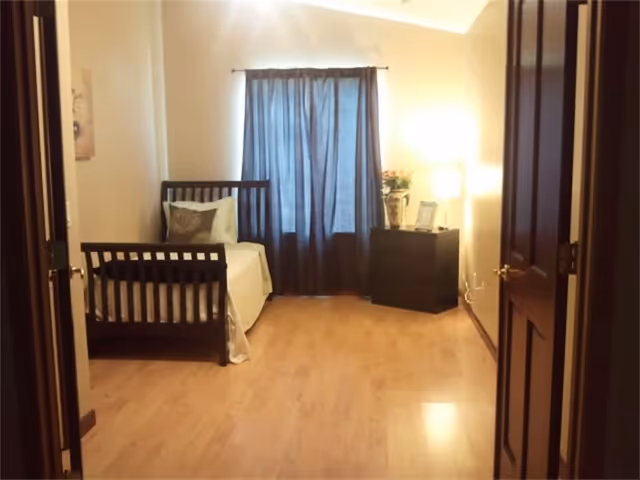 Simple furnished bedroom with a single bed to the left, dark curtains over a central window, hardwood floor and a dresser with a lamp on the right.