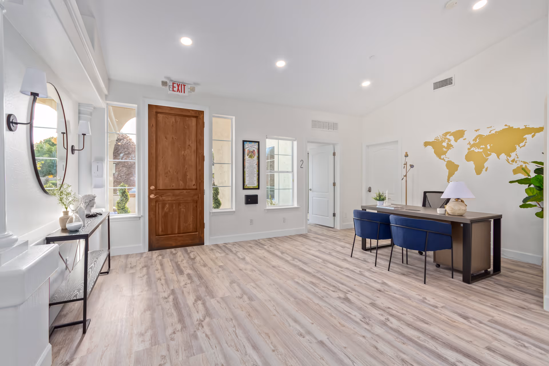 Bright reception lobby with a wooden front door, a desk with two blue chairs and a gold world map on the wall, and a console table beneath a round mirror.