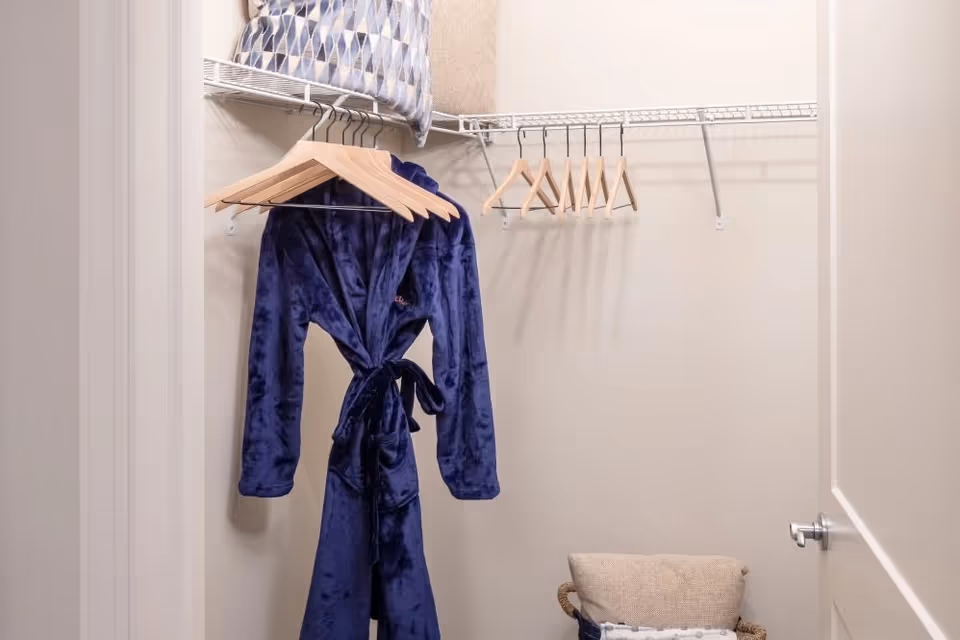 Small walk-in closet with a blue robe hanging on wooden hangers and wire shelving holding a pillow.
