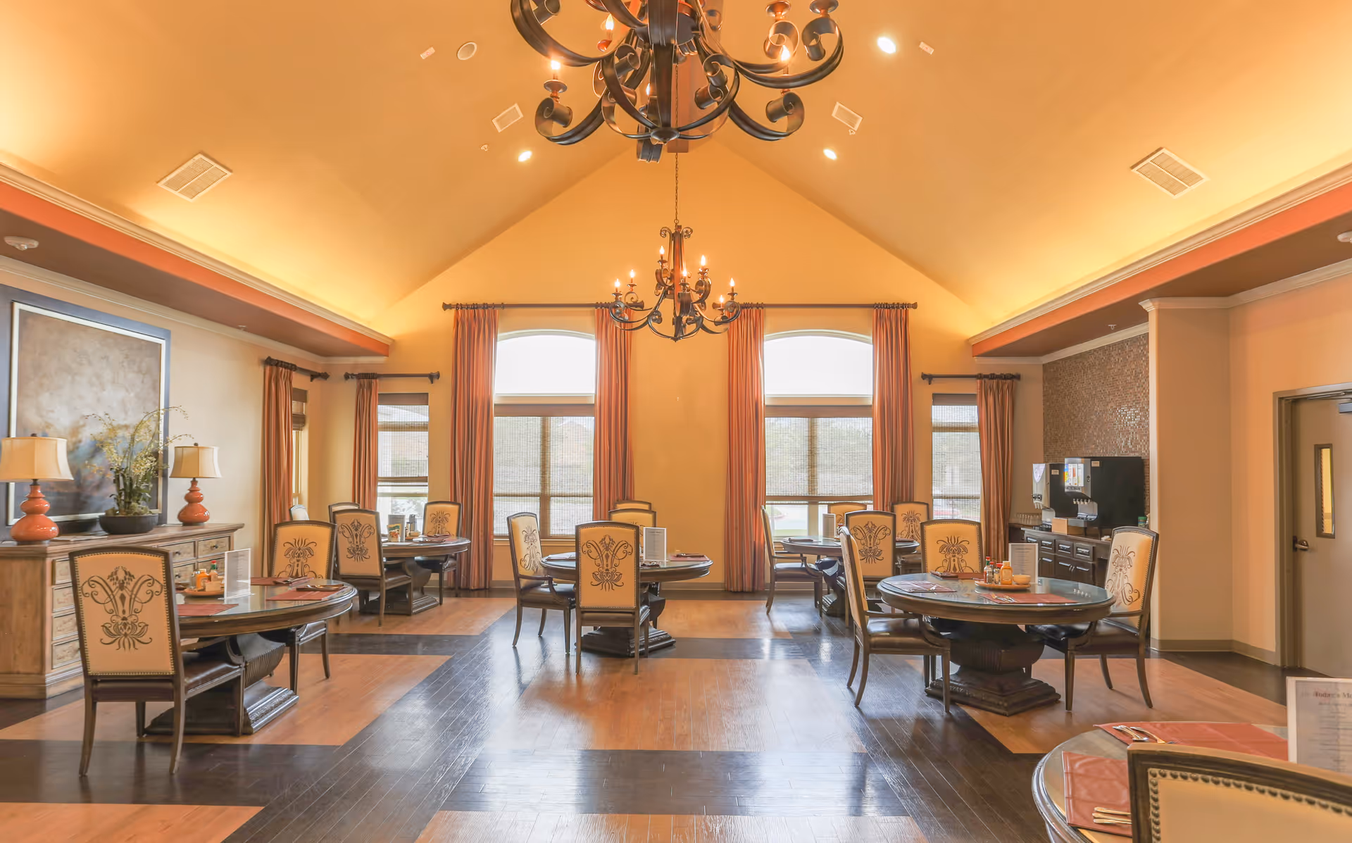 A spacious dining room with high vaulted ceilings and warm lighting. The room features several round wooden tables with upholstered chairs arranged neatly around them. Large windows with curtains allow natural light to fill the space. There are decorative elements such as table settings, lamps, and plants on a sideboard along one wall. A beverage station is visible in the corner near a door.