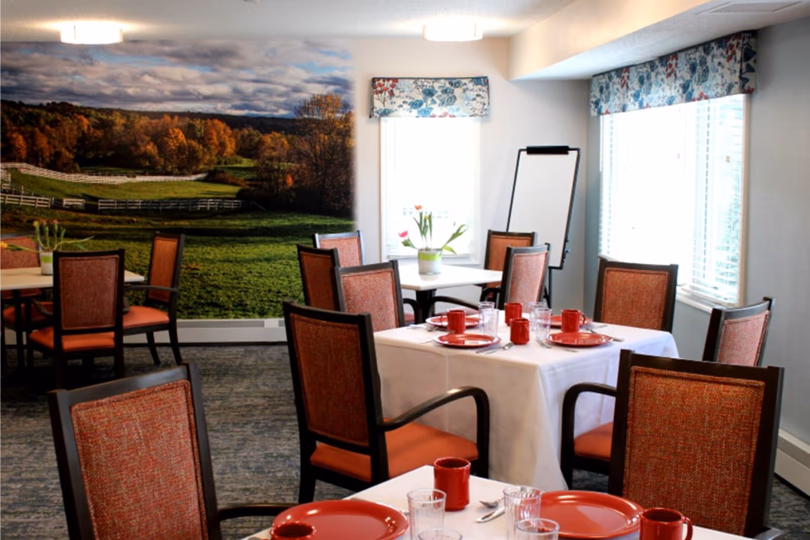 Dining room with tables set with red dishes and glasses, upholstered chairs, a large countryside mural on the wall, and windows with floral valances.