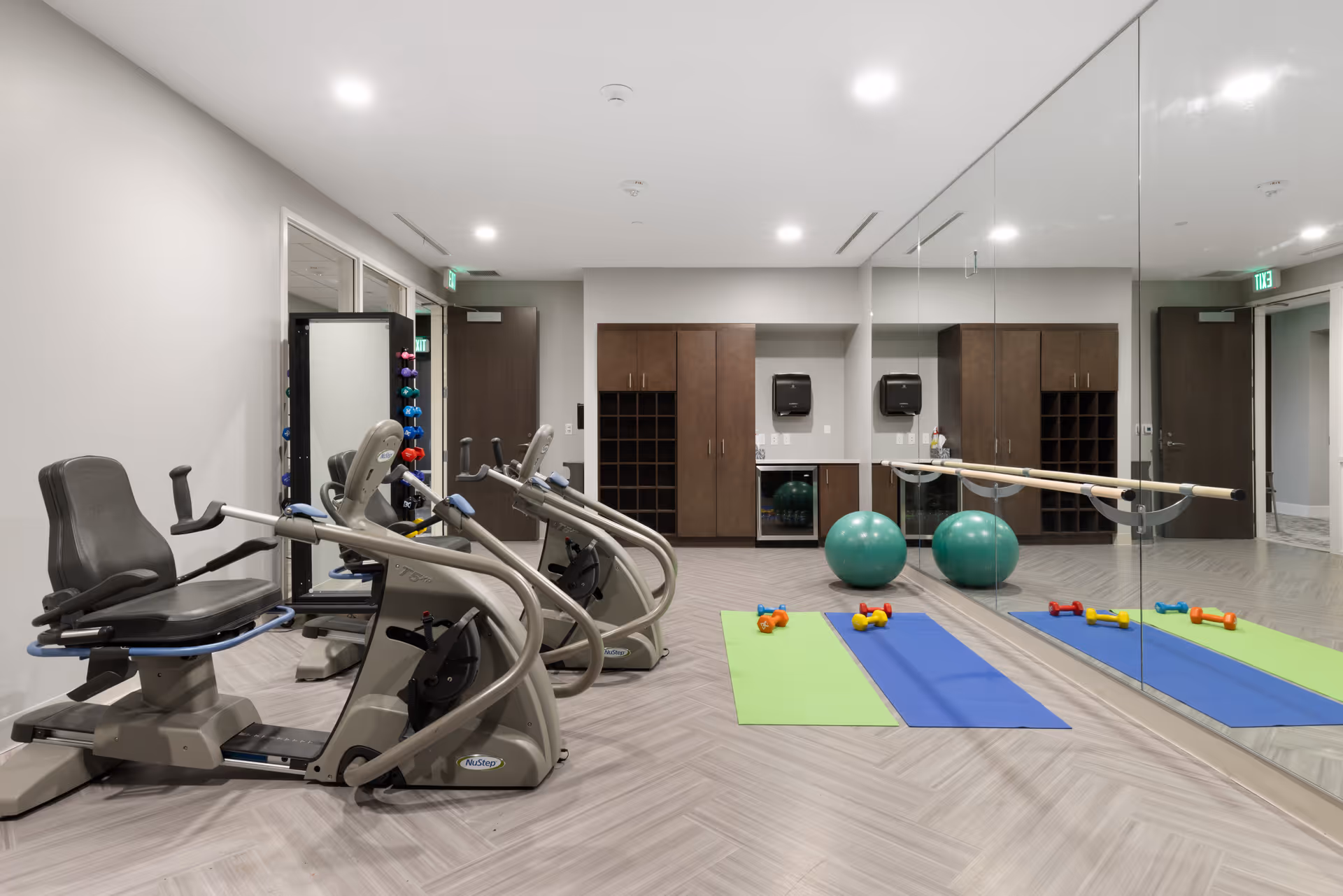 A bright fitness room with recumbent exercise bikes, yoga mats, small dumbbells, stability balls, a mirrored wall and a barre.