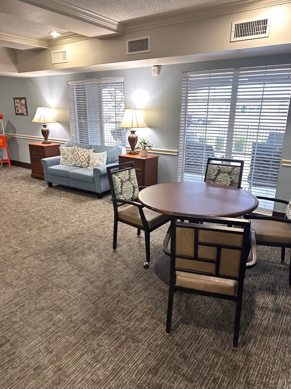 A cozy common area with a round wooden table surrounded by four chairs with patterned upholstery. Behind the table, there is a light blue sofa with decorative pillows, flanked by two wooden side tables each holding a lamp. The room has large windows with blinds letting in natural light, and the floor is carpeted in a neutral tone.
