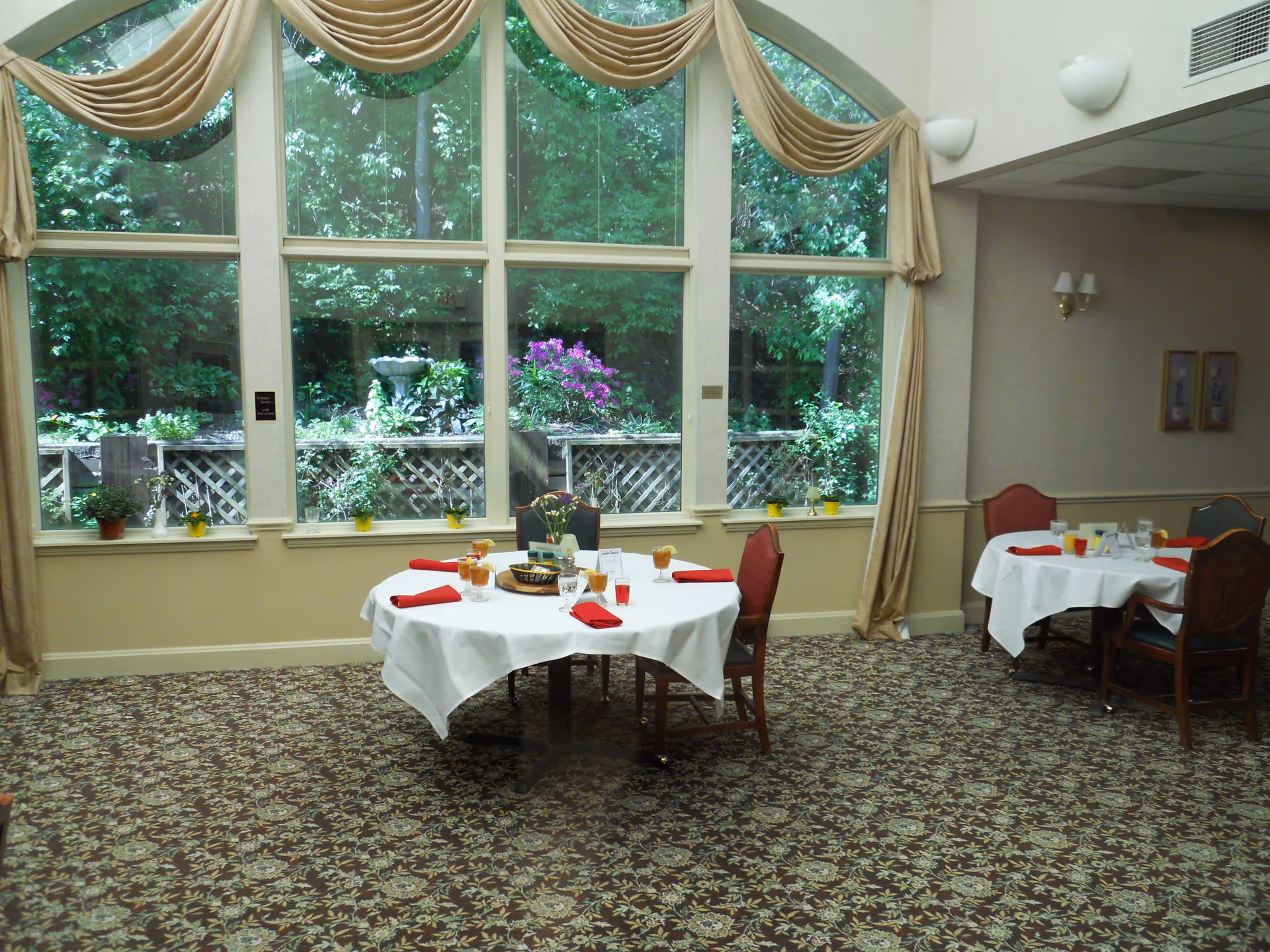 Dining room with round tables covered in white tablecloths, set with glasses, plates, and red napkins. Large windows with beige curtains reveal a garden with green trees and flowering plants outside. The room has patterned carpet and wall sconces.