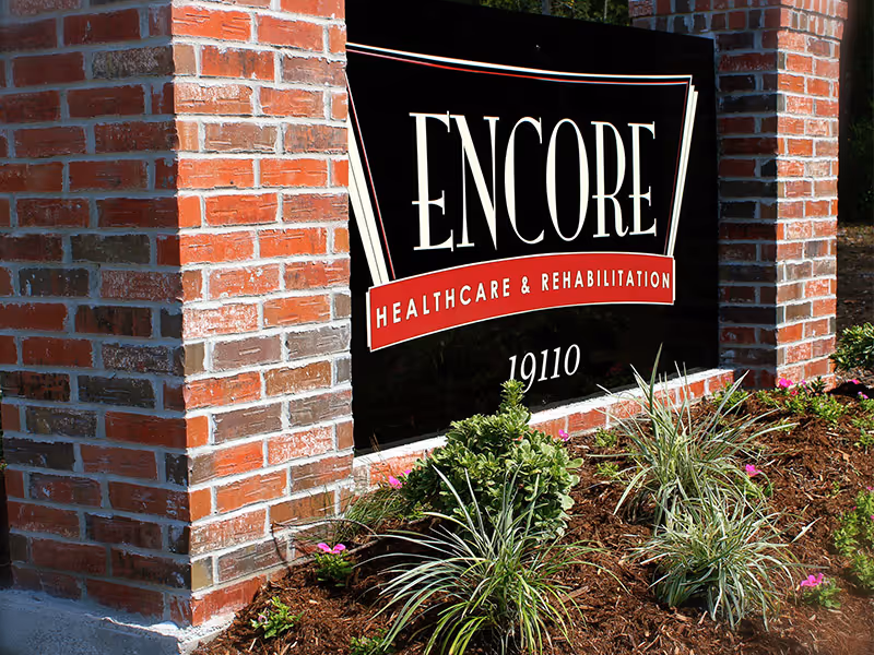 A black sign with white and red lettering that reads 'ENCORE Healthcare & Rehabilitation 19110' mounted on a brick structure surrounded by landscaping with green plants and small pink flowers.
