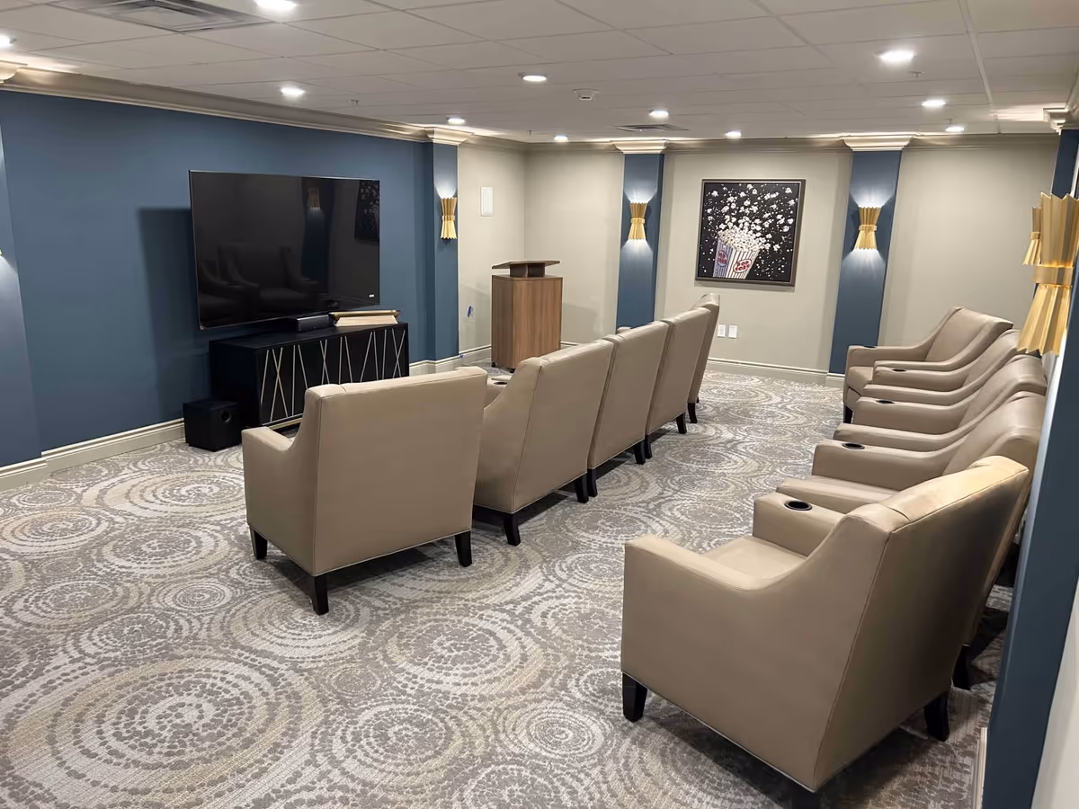 A small theater room with two rows of beige leather chairs facing a large flat-screen TV mounted on a blue accent wall. The room has patterned carpet flooring, wall sconces with gold accents, and a framed picture of popcorn on the far wall.