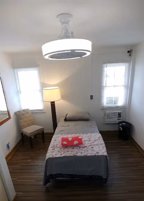 A small, simple bedroom with a single bed centered against the back wall. The bed is made with gray sheets, a pillow, and a folded pink blanket with white polka dots on top. There is a floor lamp to the left of the bed, a beige upholstered chair near the left window, and an air conditioning unit installed in the right window. The room has wooden flooring, white walls, and two windows with blinds. A modern ceiling light fixture is visible above the bed.