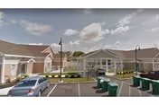 Front exterior of a single-story senior living building with a covered entrance, parking spaces, cars, and trash bins under a partly cloudy sky.