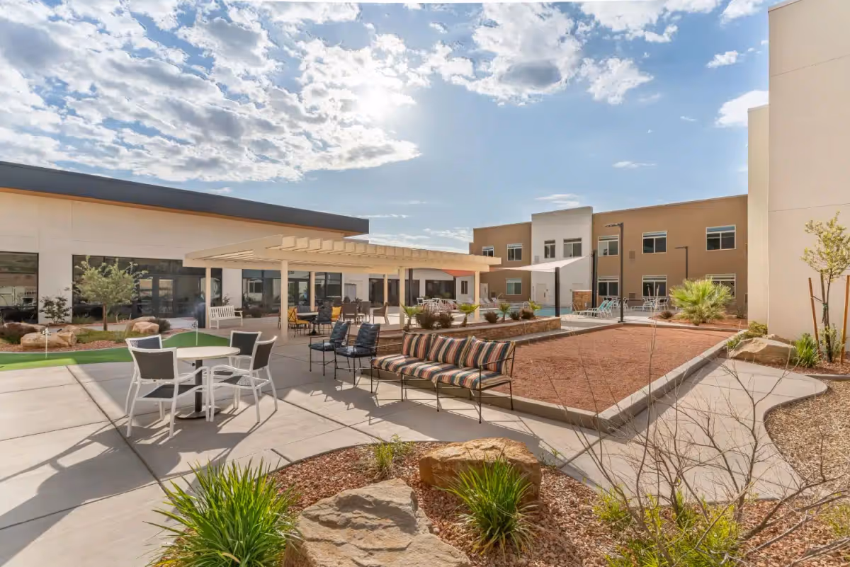 Outdoor courtyard of a senior living facility with patio seating, a pergola, landscaped planters and surrounding two‑story building under a partly cloudy sky.