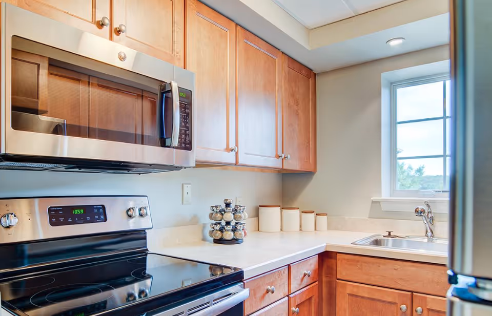 A modern kitchen with wooden cabinets, a stainless steel microwave above a black electric stove, a countertop with spice jars and canisters, and a window above a stainless steel sink.