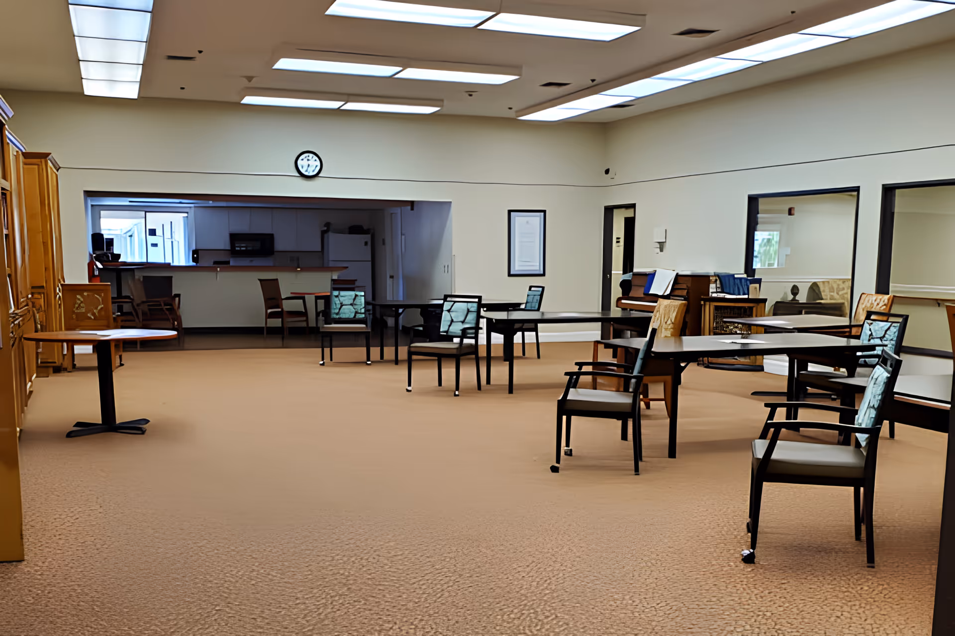 A spacious room with beige carpet and white walls featuring several tables and chairs arranged throughout. The ceiling has multiple rectangular fluorescent lights. At the back, there is a kitchen area with a refrigerator, microwave, and counter with chairs. A clock is mounted on the wall above the kitchen opening. The room has large windows on the right side and some wooden furniture, including a piano and cabinets.