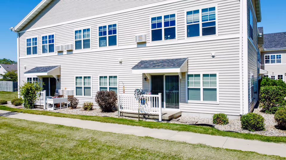 Two-story beige senior living apartment building with ground-level patios, many windows, and a sidewalk and lawn in front.