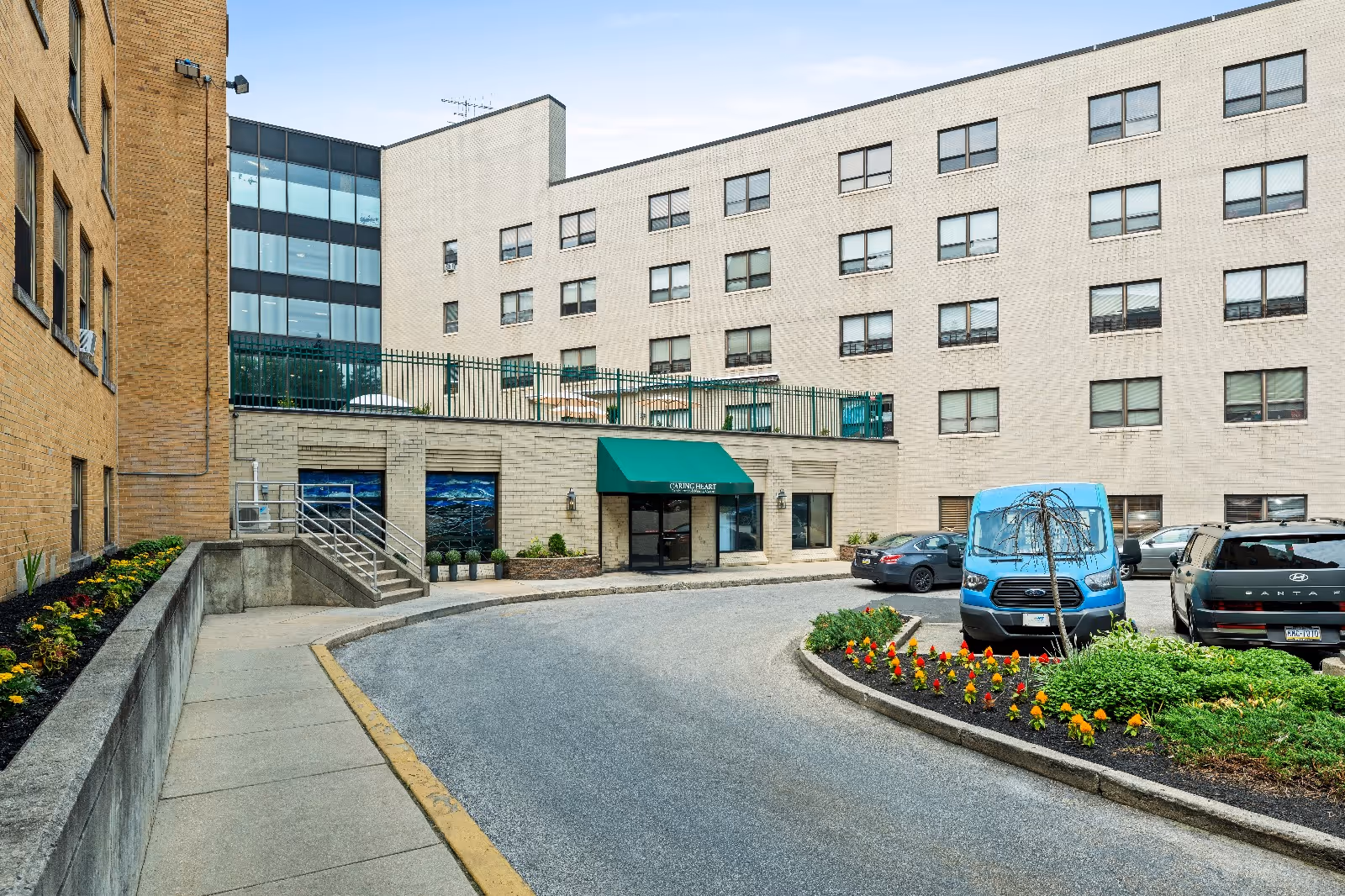 Front exterior of a multi-story nursing center with a green awning entrance, circular driveway, parked vehicles, and flowerbeds.
