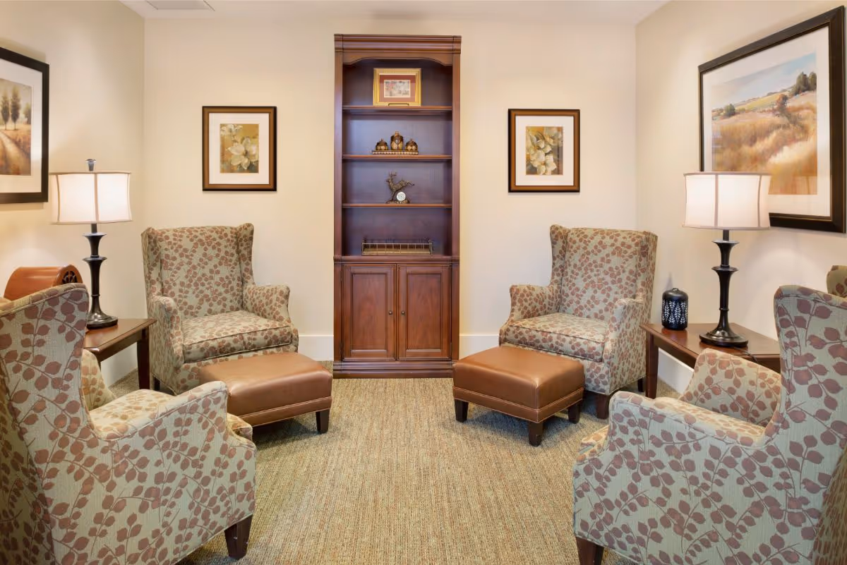 Cozy sitting room with four patterned armchairs, two ottomans, side tables with lamps, framed artwork, and a wooden shelving unit against the back wall.