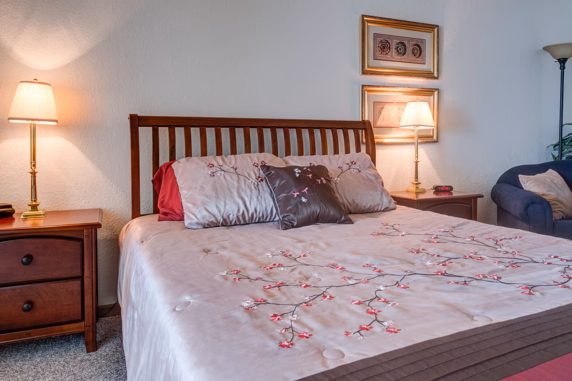 A neatly made bed with a wooden headboard, adorned with a beige bedspread featuring a floral pattern and several pillows. On either side of the bed are wooden nightstands with lamps. Two framed pictures hang on the wall above the right nightstand. A blue armchair with a pillow is visible in the corner of the room.
